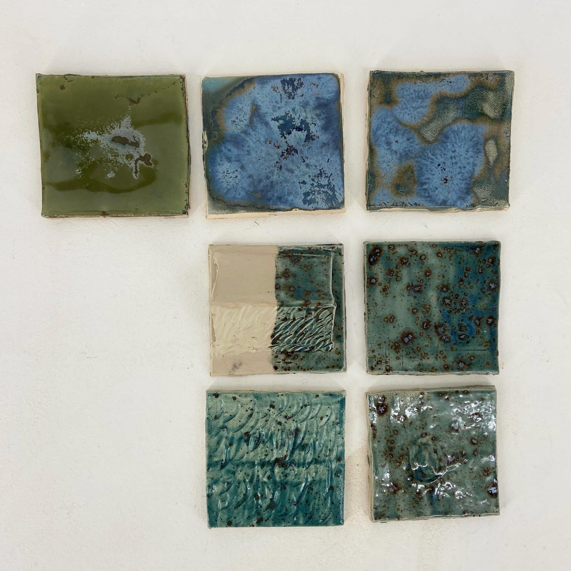 Zeven glazuurtesttegels in blauw, groen en turquoise op een witte ondergrond